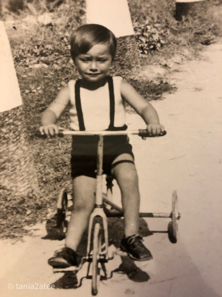 Black and white photo of a little boy on a tricycle. He is wearing black shoes and a black and white shorts and top. 